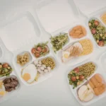 Potes de vidro com divisórias contendo porções equilibradas de proteínas, carboidratos e vegetais.