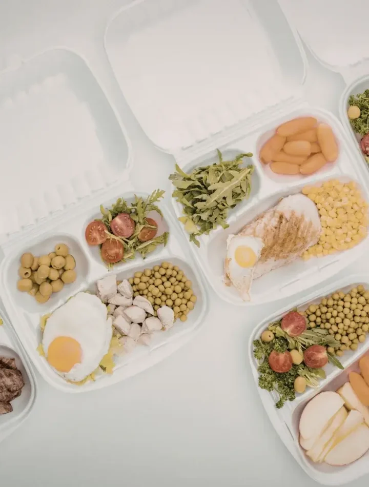 Potes de vidro com divisórias contendo porções equilibradas de proteínas, carboidratos e vegetais.